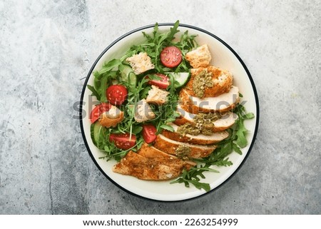Fresh salad with grilled chicken fillet, arugula, tomatoes, cucumber and pesto sauce, sesame seeds with olive oil in white bowl on light slate background. Healthy lunch menu. Diet food. Top view. 