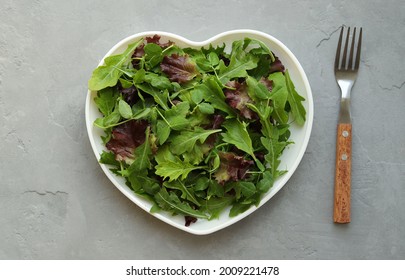 Fresh salad mixture of arugula leaves, mizuna lettuce, peas and lollo rosso in a white heart-shaped plate on a gray background. Concept: healthy food, top view. - Shutterstock ID 2009221478