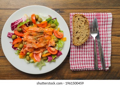 Fresh Salad With Smoked Salmon, Green Olives, Tomato, Lettuce On A White Plate. Fork, Knife And Slice Of Bread On A Red Towel. Top Down View