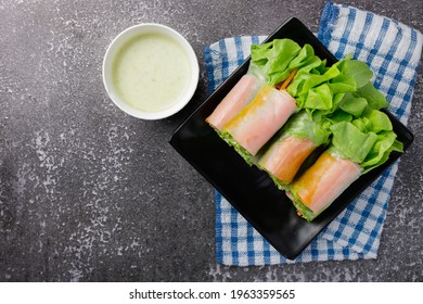 Fresh Salad Spring Roll There Are Many Kinds Of Vegetable, Lettuce, Carrot, Onion, And Crab Stick On Black Dish With Sauce In White Mini Bowl On Gray Stone Background. Top View Photo.