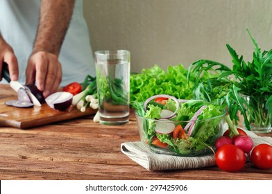 Fresh Salad Of Summer Vegetables In A Deep Bowl Of Glass. Arugula, Lettuce, Radishes, Onions, Cherry Tomatoes. In The Background Male Hand Sliced Onions On Cutting Board. Healthy Lifestyle. Copy Space