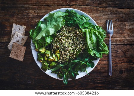 Fresh salad of vegetables and greens on a plate on a wooden background. Salad consists of Sprouted buckwheat grains, Finely chopped dill, zucchini, avocado, lettuce, spinach, Arugula and basil leaves