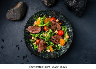 Fresh salads with beef steak, tomatoes, green leaves, arugula, tangerines and hazelnuts on a dark background. Healthy food, clean eating. Salad beef steaks, lettuce, arugula. Top view.