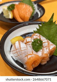 Fresh salmon sashimi slices served with shiso leaf, lemon, and garnish on a ceramic plate. A beautiful Japanese dish highlighting raw fish artistry and fine dining