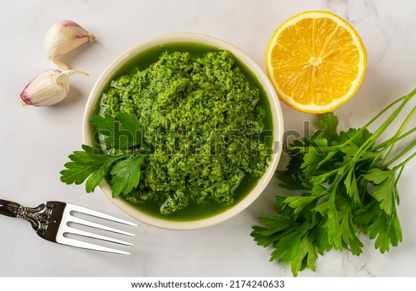Fresh salsa verde in a bowl. Chimichurri dipping\
sauce from fresh parsley, garlic cloves, olive oil and lemon juice.\
Green sauce with fresh herb and spices. Healthy condiment recipe.\
Top view.