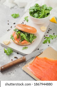 Fresh Sandwich With Bagel And Salmon, Cream Cheese And Wild Rocket On White Board With Smoked Salmon Pack And Knife On Light Background.