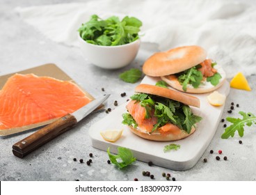 Fresh Sandwiches With Bagel And Salmon, Cream Cheese And Wild Rocket On White Board With Smoked Salmon Pack And Knife On Light Background.