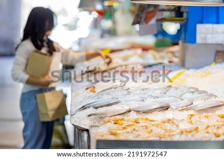 Fresh seafood on an ice counter in seafood zone at the supermarket. A counter with various seafood and Blurred woman holding brown paper bag, standing near seafood stall in background.  