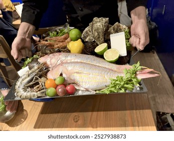 Fresh seafood platter with red snapper, prawns, oysters, lemons, and limes on a black tray - Powered by Shutterstock