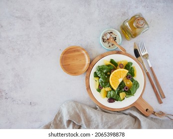 Fresh spinach salad with oranges and black olives, served on a white plate on a wooden board. Olive oil, salt and pepper for dressing. Top view on light background.