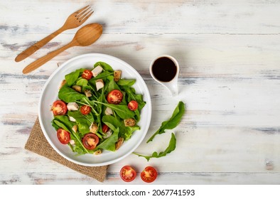 Fresh spinach salad with tomato, small pieces of herb roasted chicken in white ceramic dish on brown sackcloth, together with balsamic vinegar and utensil on white wooded table. Concept for healthy.