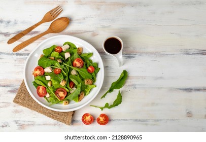 Fresh spinach salad with tomato, small pieces of herb roasted chicken in white ceramic dish on brown sackcloth, together with balsamic vinegar and utensil on white wooded table. Concept for healthy.