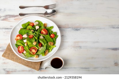 Fresh spinach salad with tomato, small pieces of herb roasted chicken in white ceramic dish on brown sack cloth, together with balsamic vinegar and metal utensil on white wooded table.