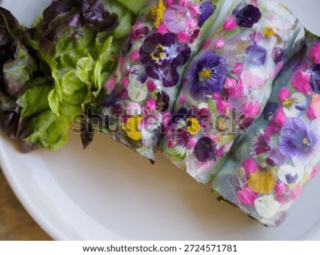 Fresh spring rolls wrapped in translucent rice paper and decorated with colorful edible flowers, served alongside leafy greens on a white plate. The image showcases creative food presentation, natural ingredients, and artistic plating, representing modern healthy cuisine and gourmet vegetarian dishes.