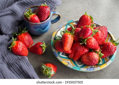 A Lot Of Fresh Strawberries On A Table. Close Up Photo Of Blue Ceramic Plate And Cup, Juicy Berries And Navy Blue Kitchen Towel. Grey Concrete Background. 