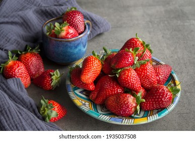 A Lot Of Fresh Strawberries On A Table. Close Up Photo Of Blue Ceramic Plate And Cup, Juicy Berries And Navy Blue Kitchen Towel. Grey Concrete Background. 