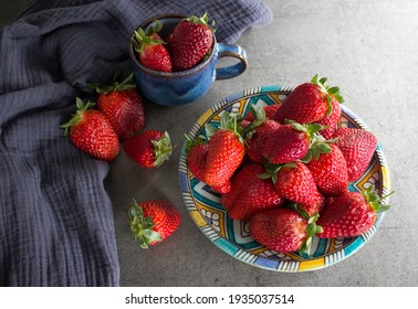 A Lot Of Fresh Strawberries On A Table. Close Up Photo Of Blue Ceramic Plate And Cup, Juicy Berries And Navy Blue Kitchen Towel. Grey Concrete Background. 