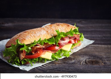Fresh submarine sandwich with ham, cheese, bacon, tomatoes, cucumbers, lettuce and onions on dark wooden background