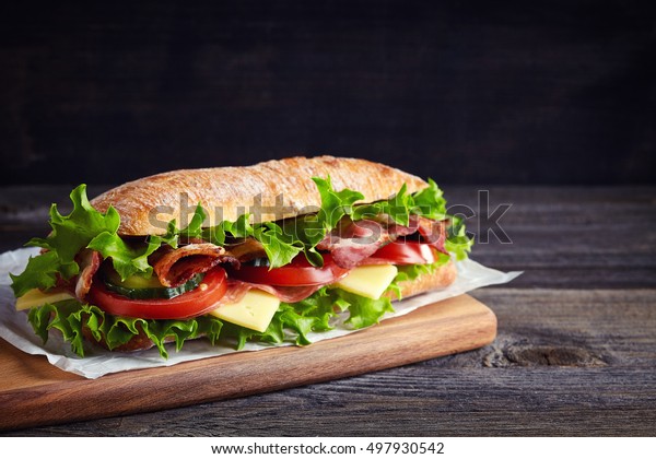 Fresh\
submarine sandwich with ham, cheese, bacon, tomatoes, cucumbers,\
lettuce and onions on wooden cutting\
board