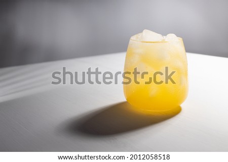 Fresh summer cold orange juice with ice in glass in sunlight with striped shadow on white table with gradient wall in modern simple abstract style.