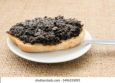 Fresh Tapenade Sandwich With Slice Of Wheat Bread In A White Sauser And Table Knife On A Burlap Table Napkin. Homemade Black Olive Paste With Anchovies, Garlic And Olive Oil. Front View.