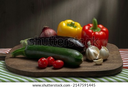 Fresh tasty colorful vegetables as eggplant, zucchini, yellow and orange paprika, red onion, garlic, cherry tomatoes for cooking of french ratatoille on brown cutting board laying on striped towels.
