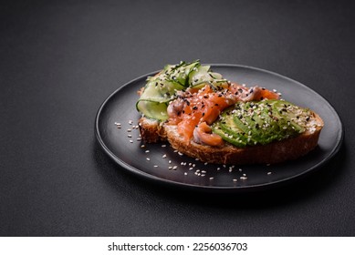 Fresh tasty sandwich with salmon, avocado and sesame and flax seeds on a dark concrete background - Shutterstock ID 2256036703