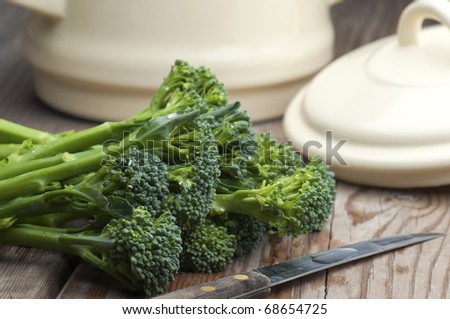 Fresh Tenderstem Broccoli Laid On A Wooden Kitchen Table With A Vegetable Knife and A Pan With Lid In The Background