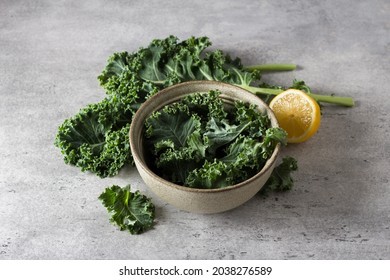 Fresh torned kale leaves in a bowl with lemon and whole kale leaves on gray textured background, top view. Cooking kale salad