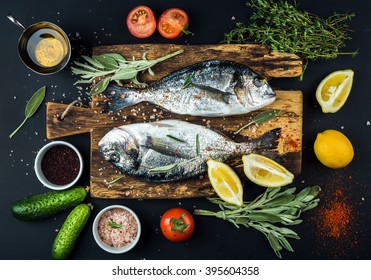 Fresh uncooked dorado or sea bream fish with lemon, herbs, oil, vegetables and spices on rustic wooden board over black backdrop, top view