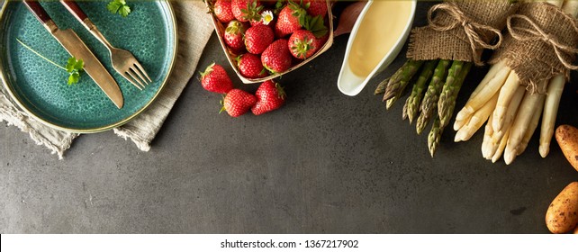 Fresh uncooked green and white asparagus, mayonnaise, potatoes and strawberries with an empty plate and utensils in panorama banner format viewed from above