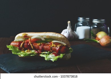 Fresh Vegan Hot Dog On The Background Of Salt, Pepper, Garlic And Bulbs. Hotdog With Soy Sausages, Fresh Salad, Cucumbers, Tomatoes And Onions