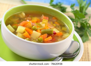 Fresh vegetable soup made of green bean, pea, carrot, potato, red bell pepper, tomato and leek in bowl with parsley in the back (Selective Focus, Focus on the vegetables one third into the soup)