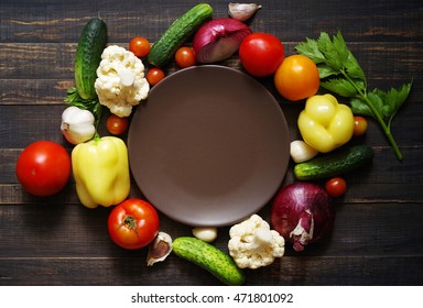 Fresh vegetables around a round brown empty plate on a wooden background of a table from above. Healthy, vegetarian, organic food  