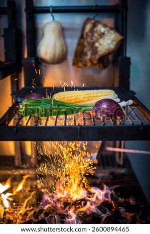 Fresh vegetables and corn are placed on a metal grill above an open flame, while sparks fly upwards, emphasizing the intensity of the heat. In the background, dried ingredients hang from the grill.