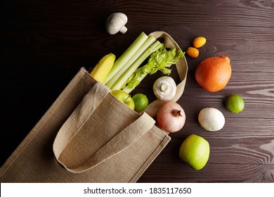 Fresh vegetables and fruits in reusable shopping bag on wooden table, top view. Healthy raw food in jute bag. Apples, citrus fruits, mushrooms, pomegranate, celery