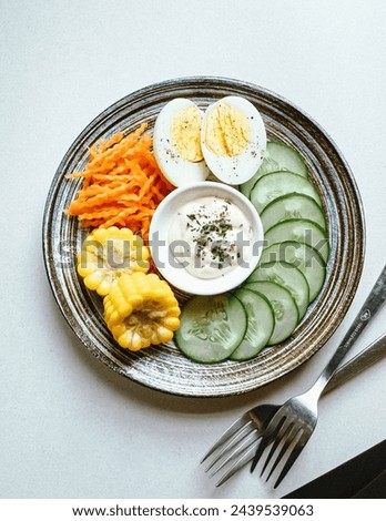Fresh vegetables with hard boiled egg. Sliced cucumber, carrots, boiled corn, and yogurt sauce. Served on a rustic round plate, next to forks and napkin. Top view