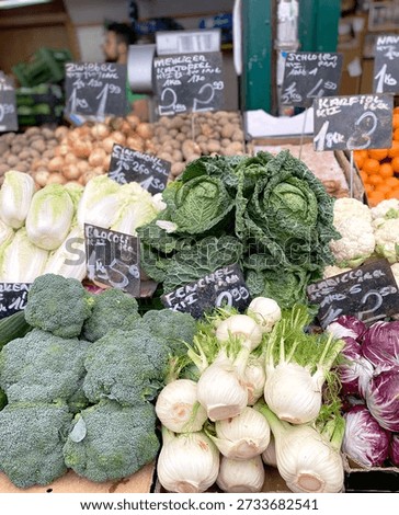 Fresh vegetables such as broccoli, cabbage, and fennel are arranged on a market stall with visible price tags on a wooden table