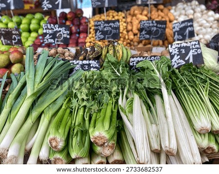 Fresh vegetables such as leeks and celery are arranged at a market stall, with colorful fruits and price tags visible in the background