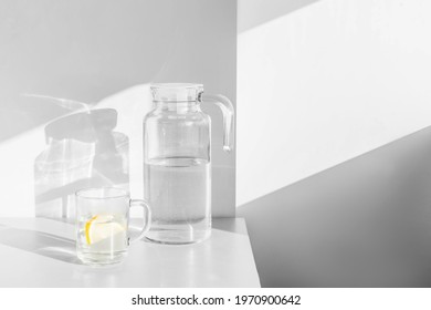 Fresh water with lemon slices in carafe and glass on white table with shadows and light. 