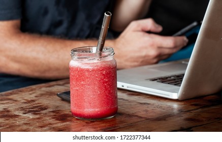 Fresh Watermelon Juice In Glass With Metal Straw On Brown Wooden Table. Laptop And Man Hand Holding Smartphone Visible On Background. Working At Cafe. Home Office 