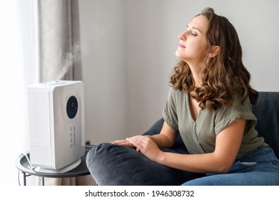 Fresh and wellness young woman using air humidifier at cozy apartment, female sits on the sofa with eyes closed and enjoys clean air spreading with modern humidifier, breaths to full chest