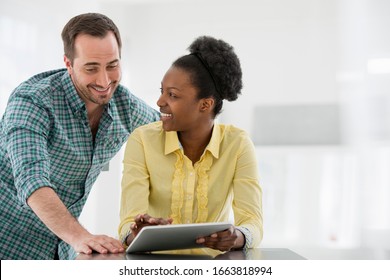 A Fresh White Building Interior, Flooded With Light. A Young Woman At A Table Using A Digital Tablet, And A Man Looking Over Her Shoulder.