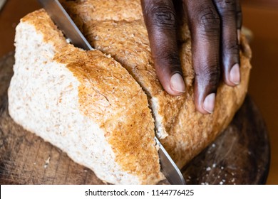 Fresh Whole Grain Bread On Kitchen Table Or Wooden Bread Board With A Black African American Man Holding Bread Knife And Cutting A Slice From Loaf For Healthy Eating And Traditional Bakery Concept