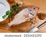 Fresh whole raw alfonsino fish prepared for cooking on wooden kitchen table accompanied by green parsley, garlic cloves, onion, and olive oil