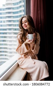 Fresh young woman in pink tender bathrobe drink tea,  looking out the window. 
