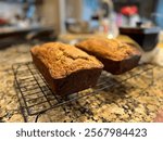 “Two freshly baked banana bread loaves cool on a wire rack in a warm kitchen. The golden-brown crust and moist texture highlight their homemade charm, set on a granite countertop in a cozy baking scen