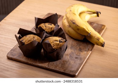 Freshly baked banana chocolate chip muffins wrapped in brown parchment paper, placed on a wooden cutting board alongside a bunch of ripe bananas. The rustic setup on a wooden table - Powered by Shutterstock