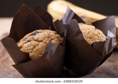 Freshly baked banana chocolate chip muffins wrapped in brown parchment paper, placed on a wooden cutting board alongside a bunch of ripe bananas. The rustic setup on a wooden table - Powered by Shutterstock