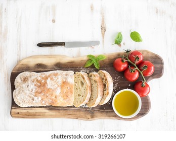 Freshly baked ciabatta bread with cherry-tomatoes, olive oil, basil and salt on walnut wood board over white background, top view, copy space